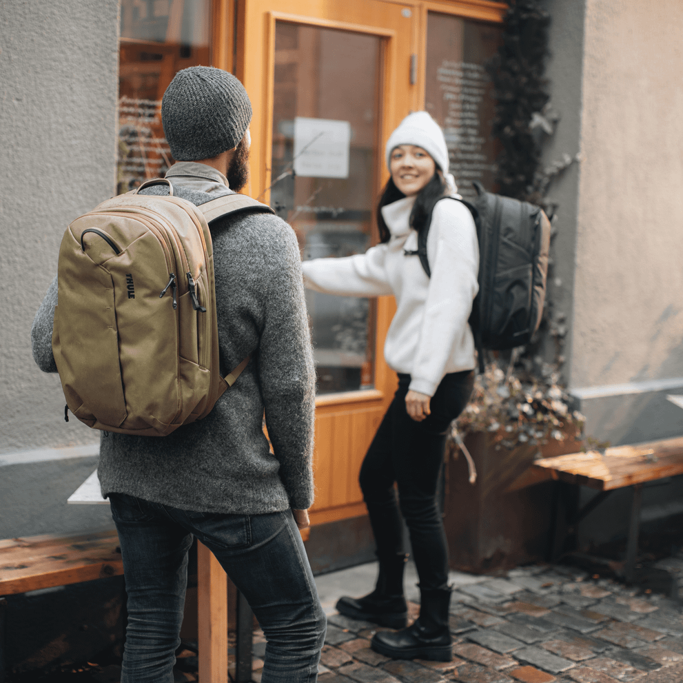 A woman in a white hat with a black Thule Aion Backpack opens the door for a man with a tan backpack.