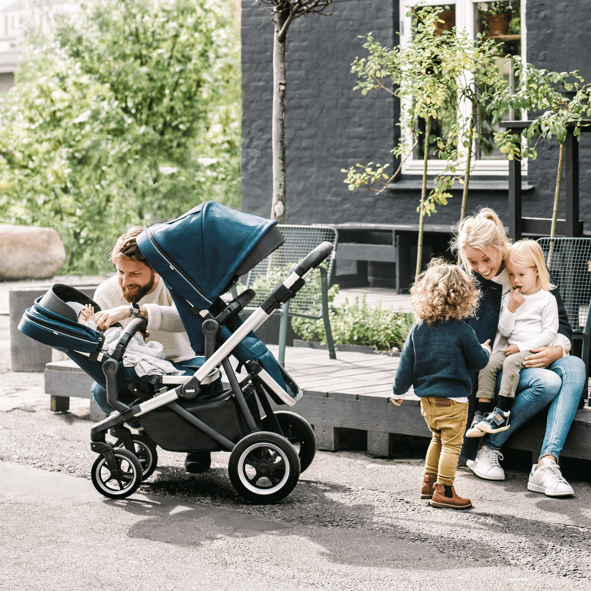 Parents sit in a cafe outside with their kids running around a stroller with a blue Thule Sleek Sibling Seat.