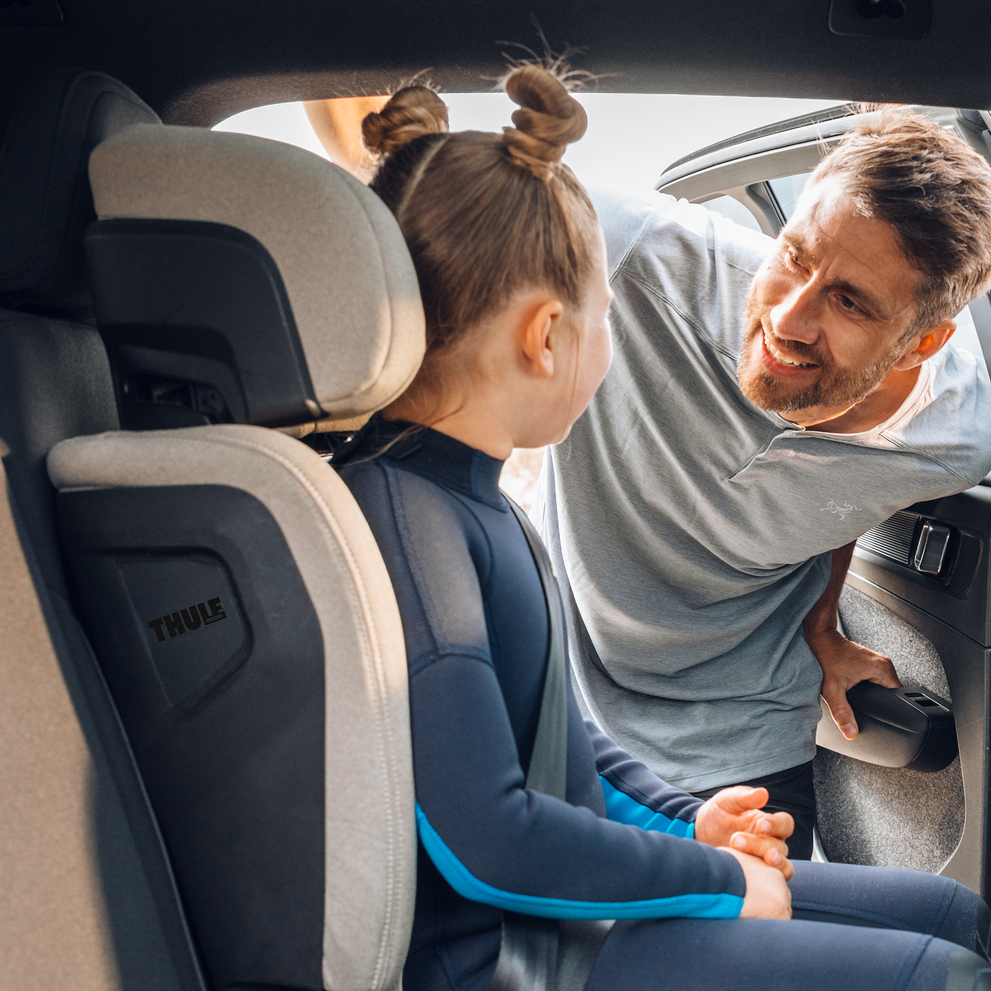 Een man die een jong meisje controleert dat in een Thule Palm hoog-achtersteun stoel zit achterin de auto.