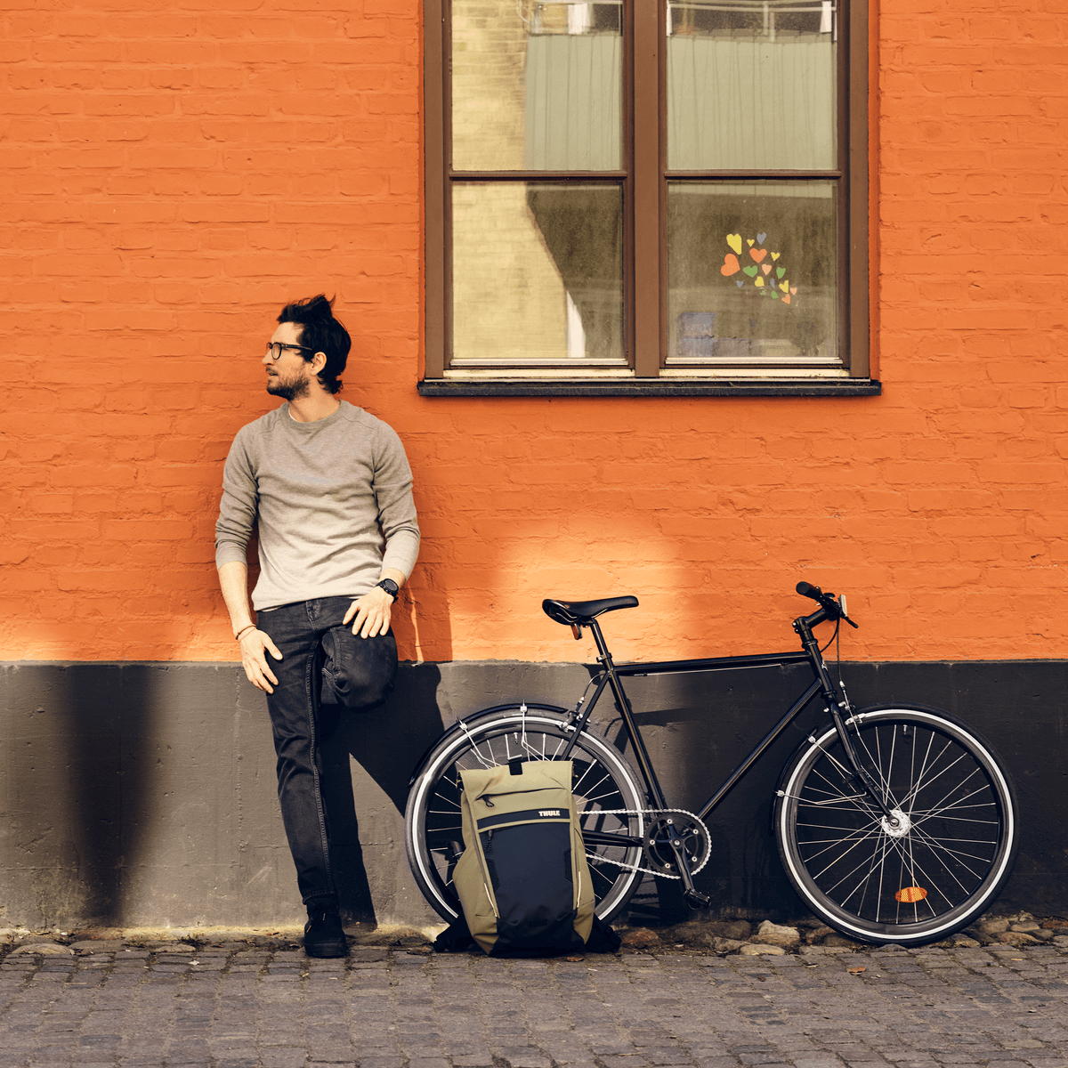 A man leans against an orange wall with a bike and a green 18L Thule Paramount commuter backpack.