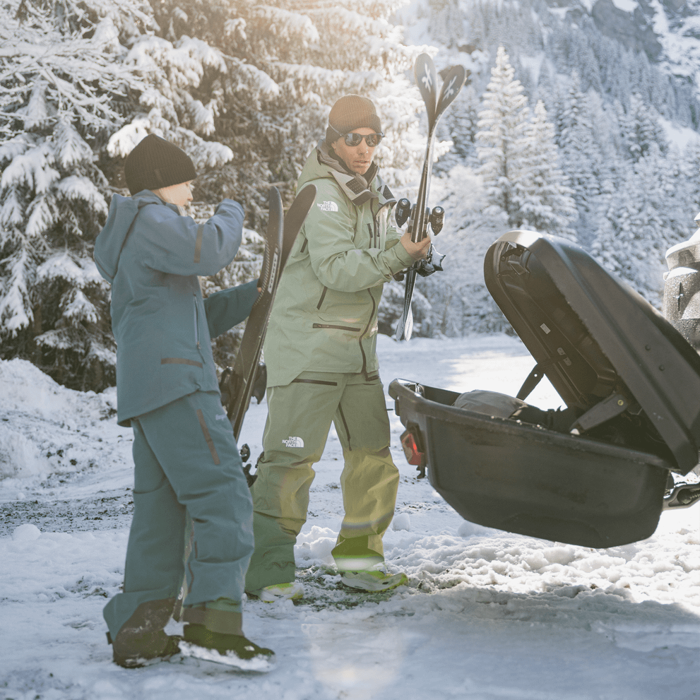 Two people dressed in ski gear, prepare to load their ski equipment into an open, Thule Arcos XL, surrounded by snow-covered trees and winter sunshine.