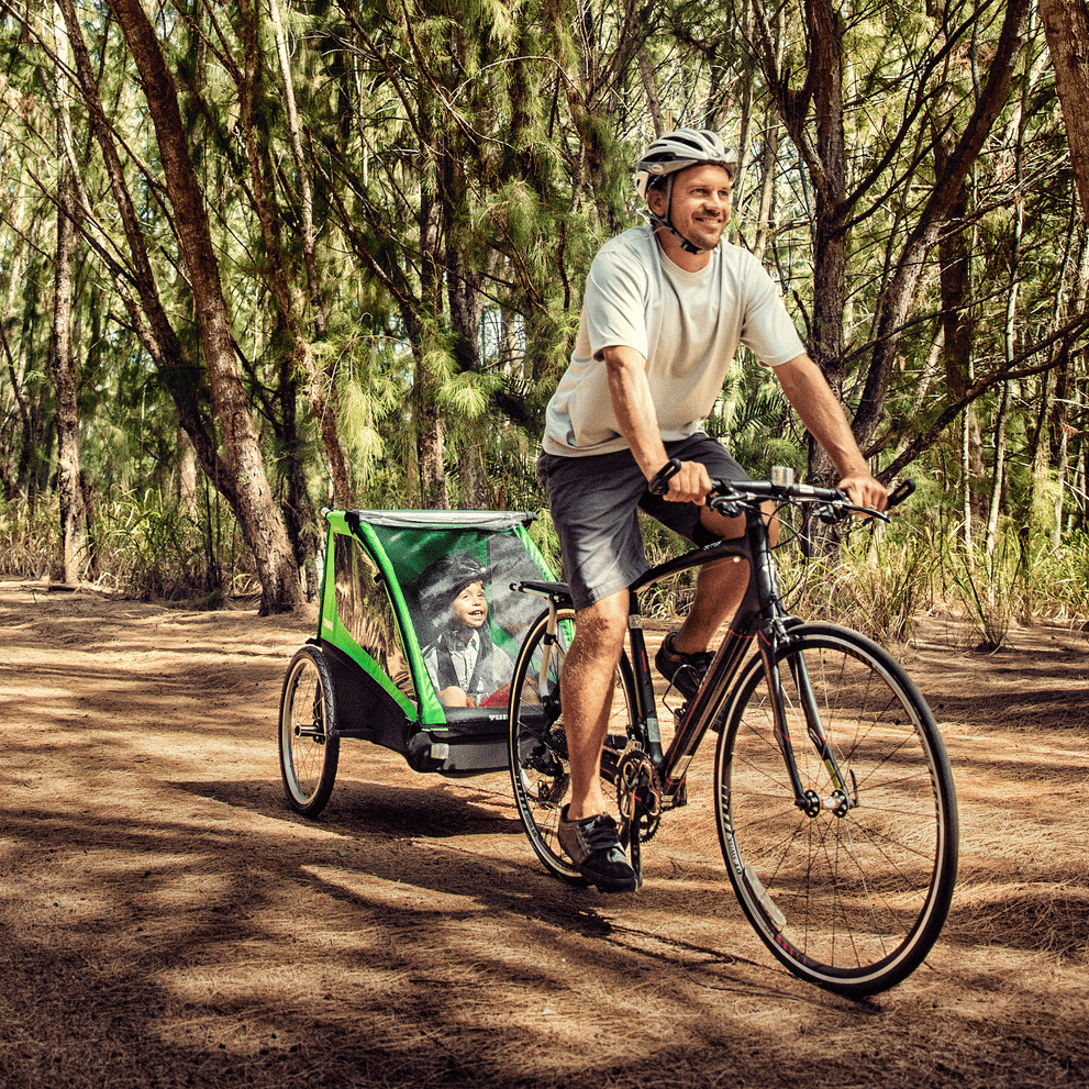 A man bikes through a forest with a green Thule Cadence bike trailer.