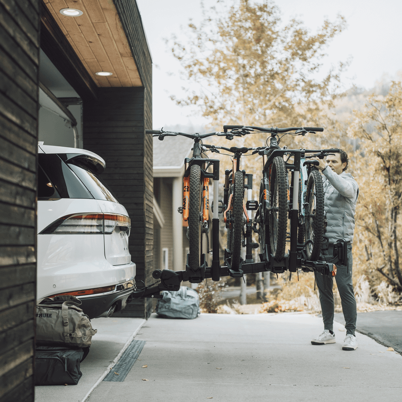 A man loads four bikes into Thule Verse hitch bike rack installed on a car parked in a garage.