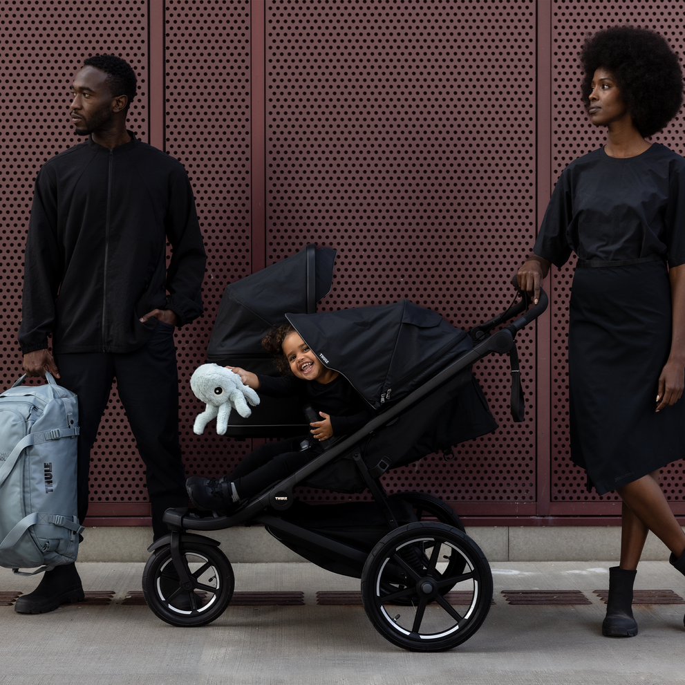 A man and a woman stand next to their black Thule Urban Glide 3 double stroller.
