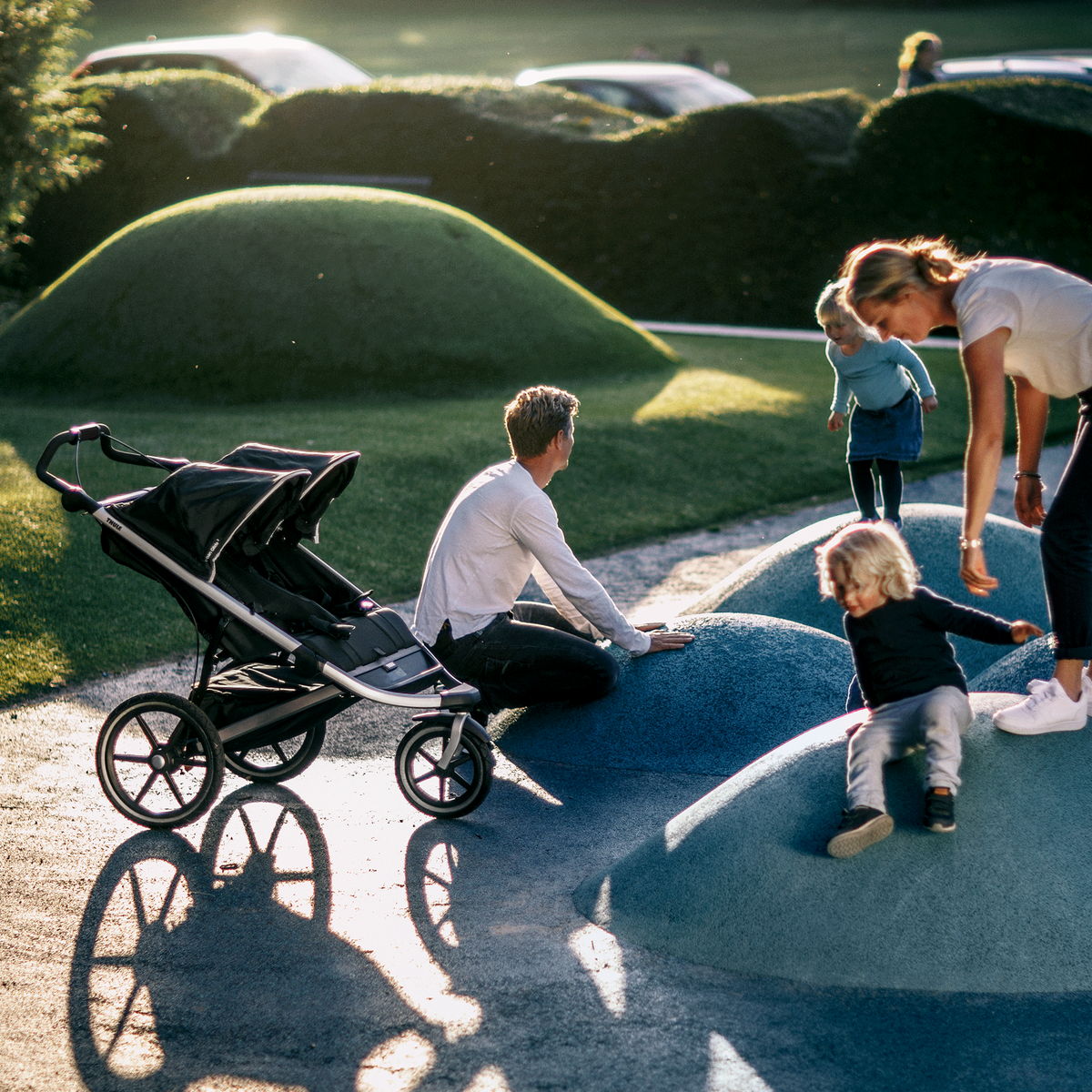A family play in a playground in the sunshine next to their black Thule Urban Glide 2 double jogging stroller.