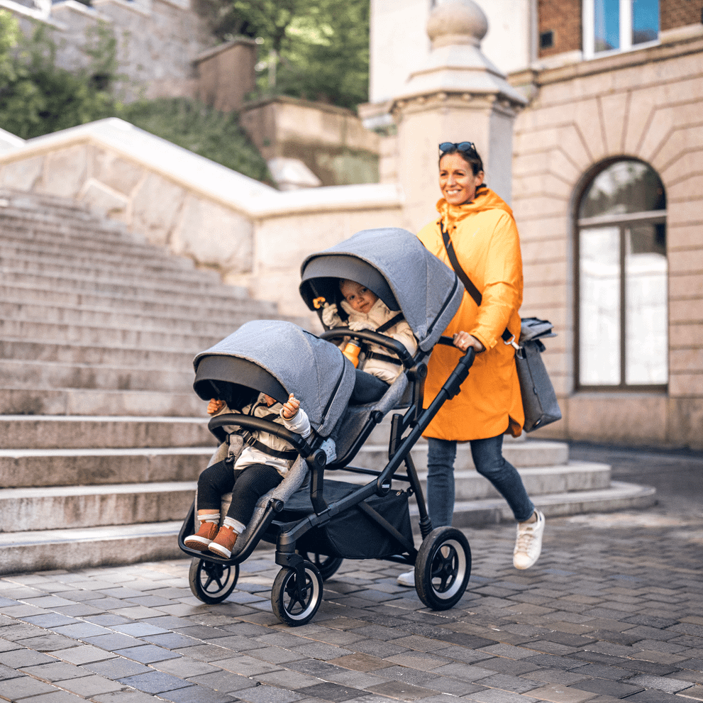 A woman in orange walks down a cobbled street with her kids in a gray Thule Sleek Sibling Seat.