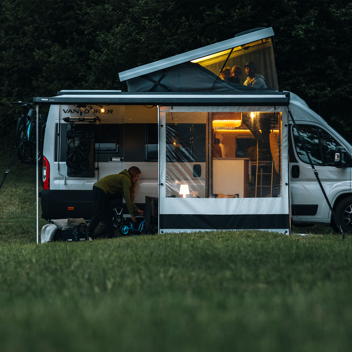 A white van at night with lights and a Thule Rain Blocker Front awning side wall.
