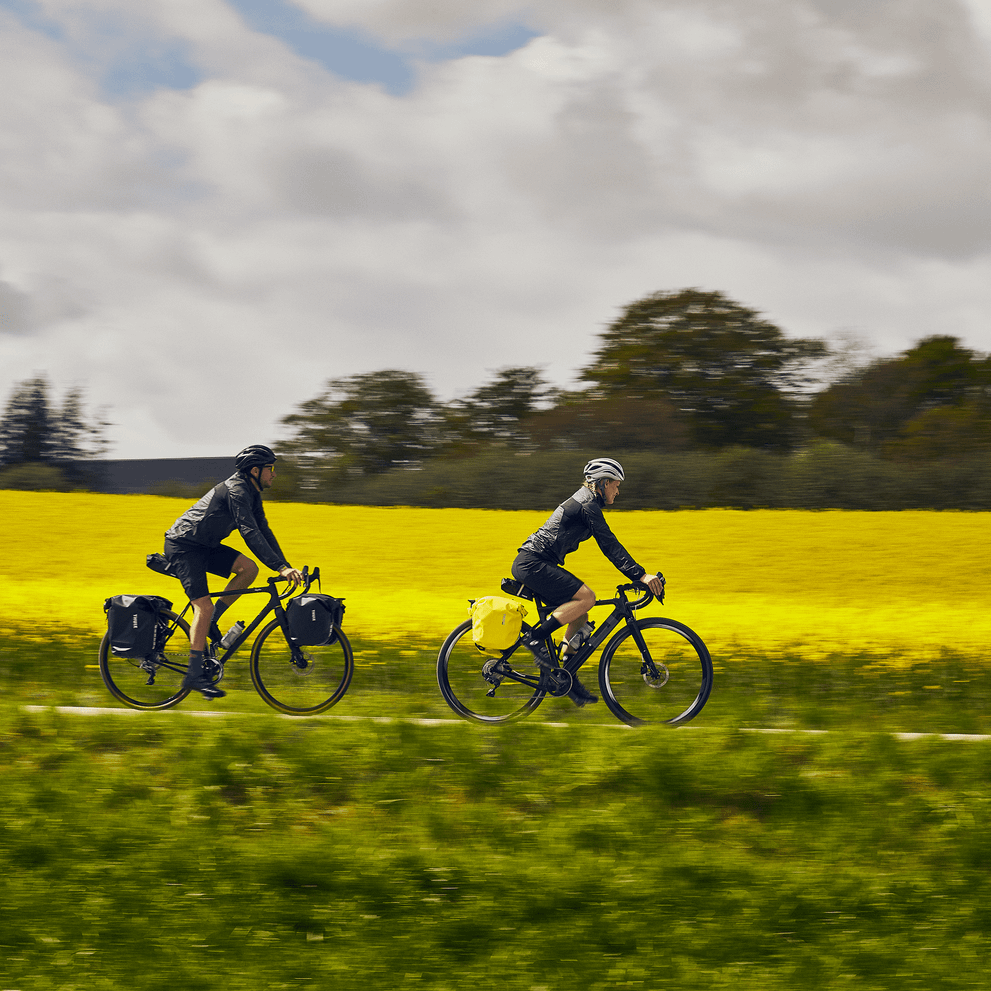 A couple bike down a country path with yellow flowers, carrying Thule Shield 25L bike bags.