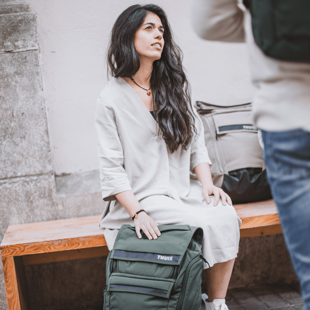 A woman sits on a bench and looks up at someone, her green Thule Paramount backpack leaning against her.