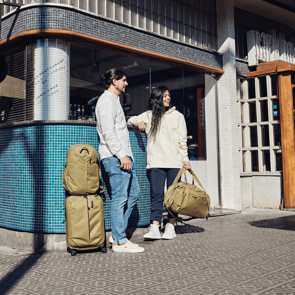 A couple stand in the sun by a restaurant with tan bags including a Thule Aion carry on spinner.