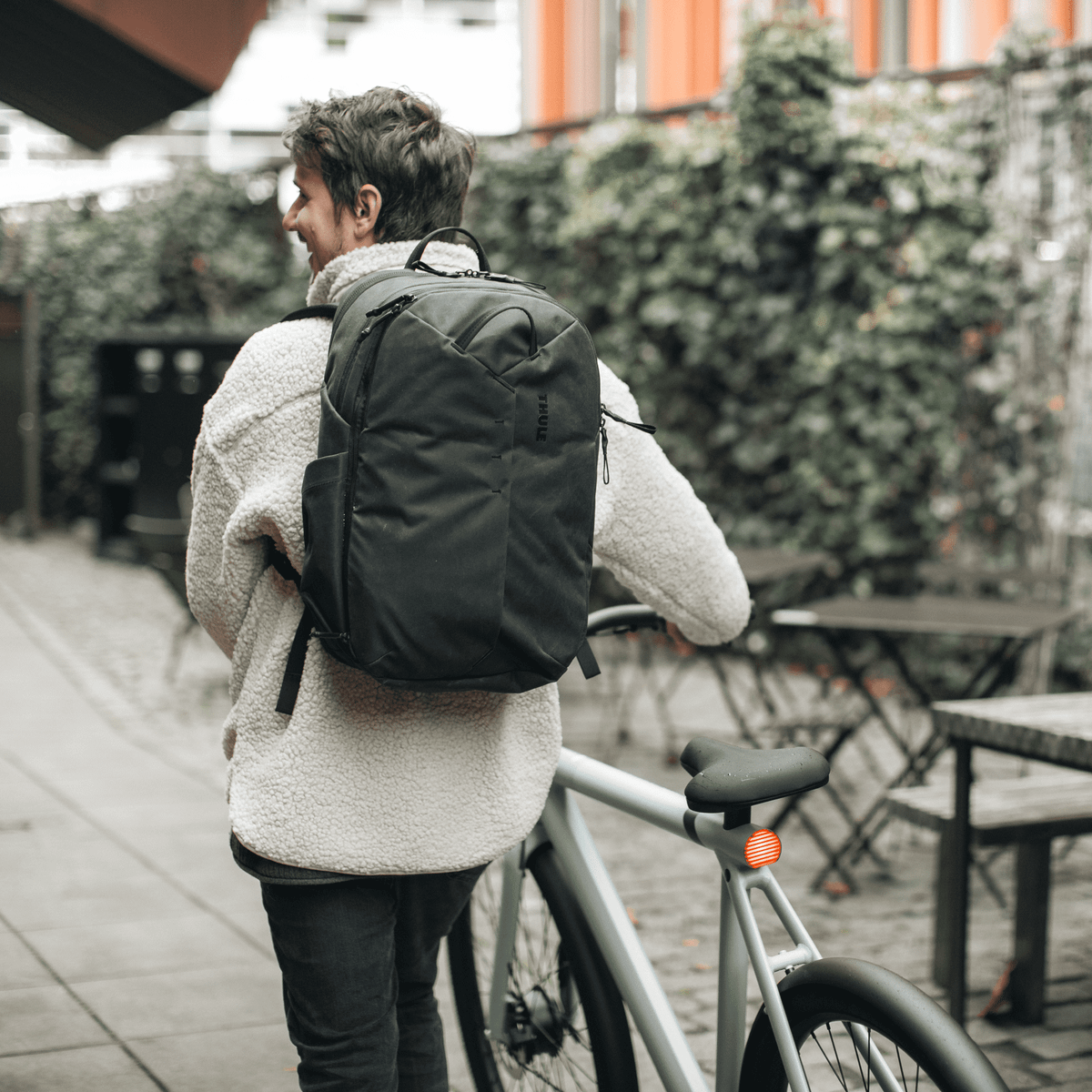 A man in a fleece walks down a street with a bike and a black Thule Aion Backpack.