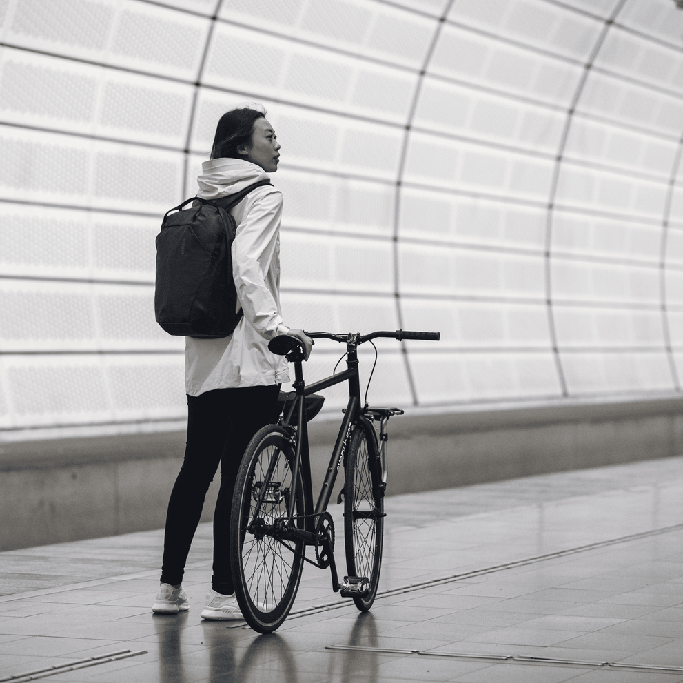 A woman holds a black bike in a train station, with a white jacket and a black Thule Tact backpack.