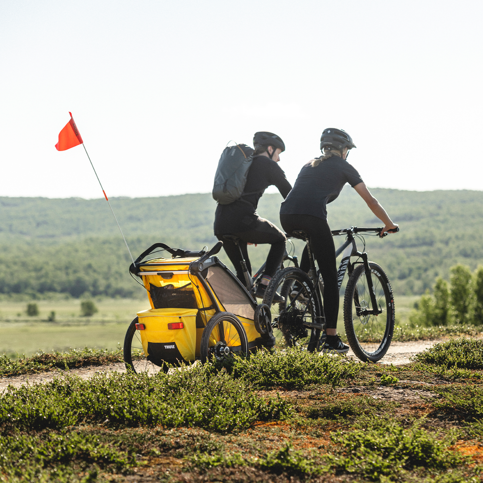 Two cyclists bike through the countryside, one with a yellow Thule Chariot Sport kids' bike trailer.