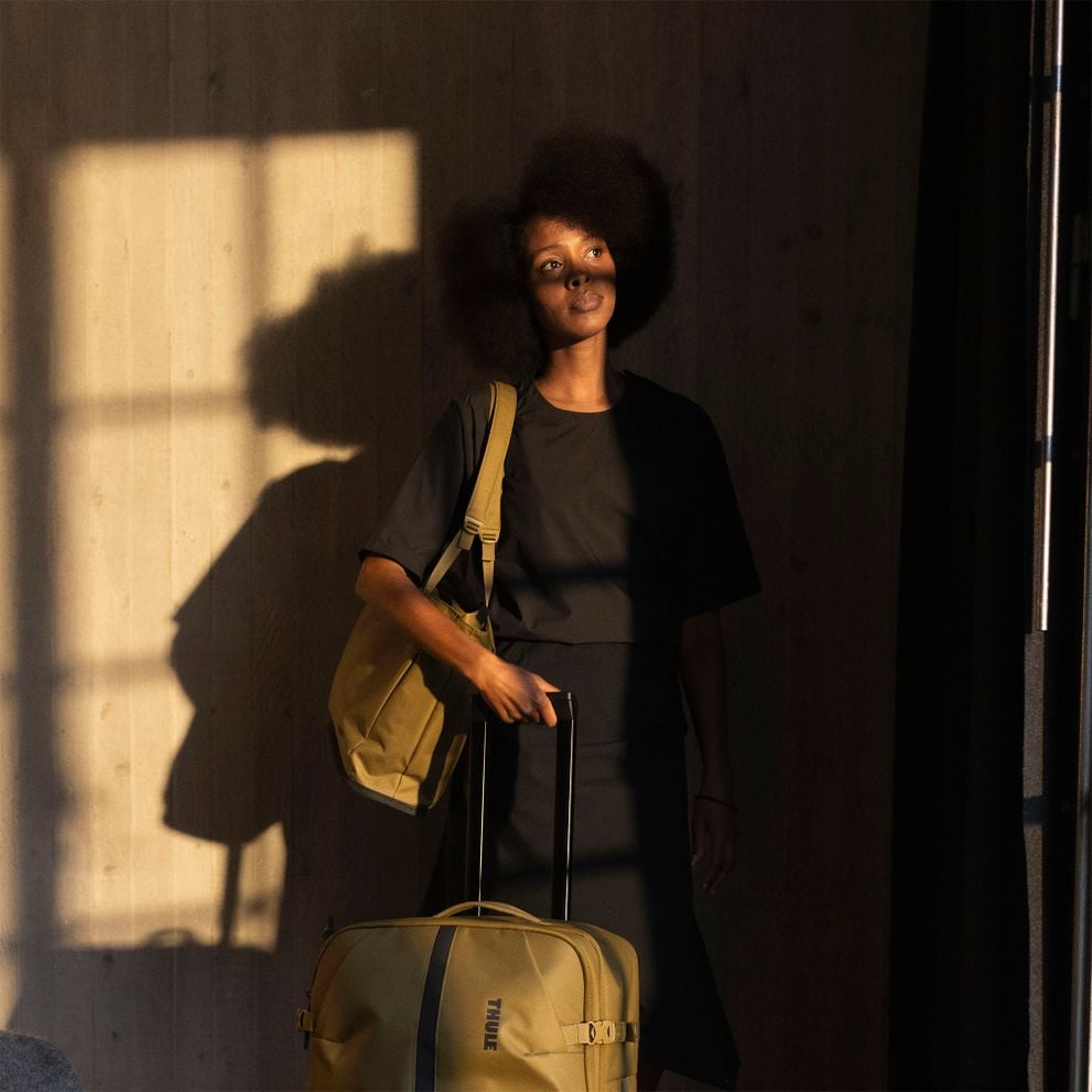 Mujer junto a una ventana con una maleta rodante Thule Aion bajo la cálida luz del sol.