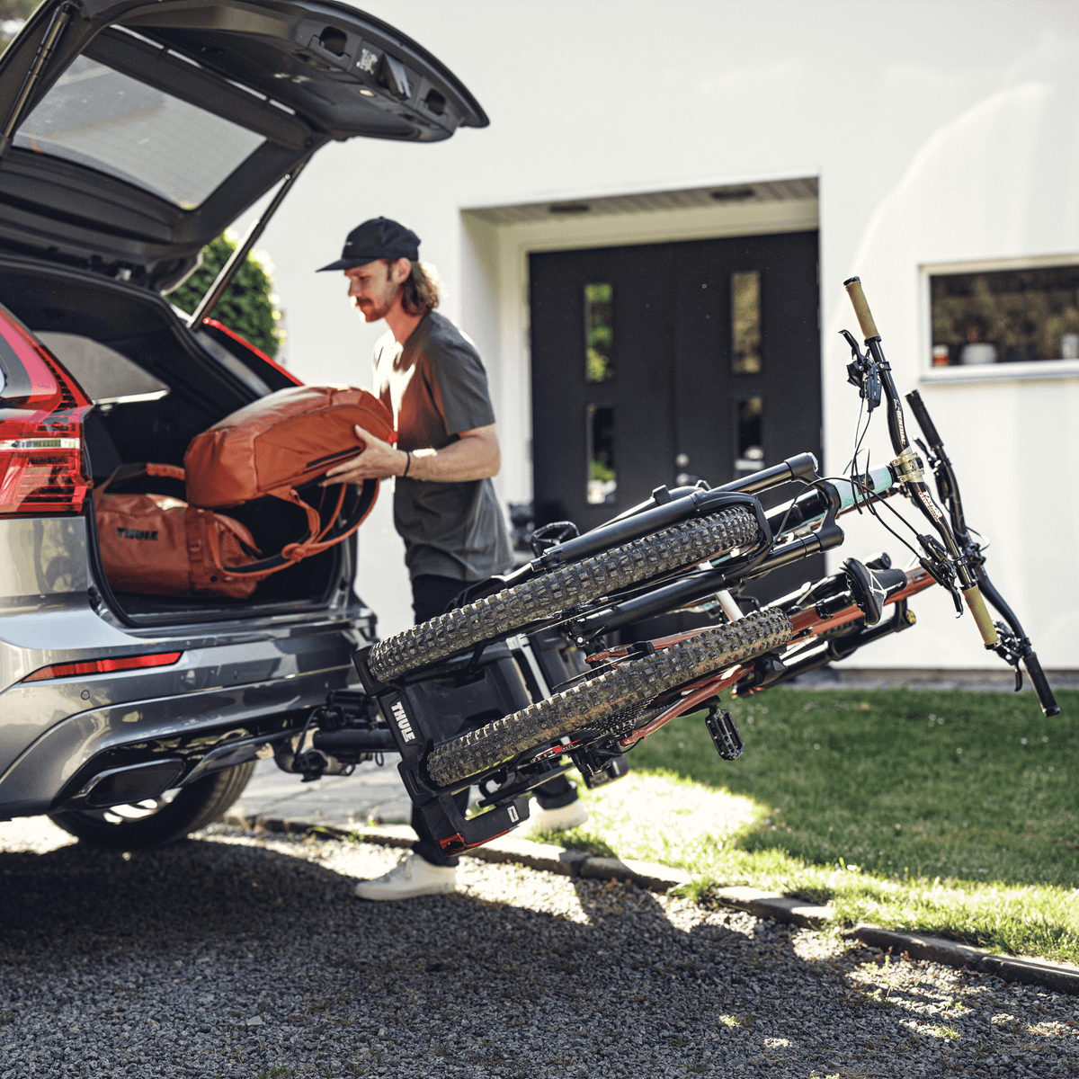 A man is loading bags in the trunk of his car, where a bike carrier is mounted on the towbar.