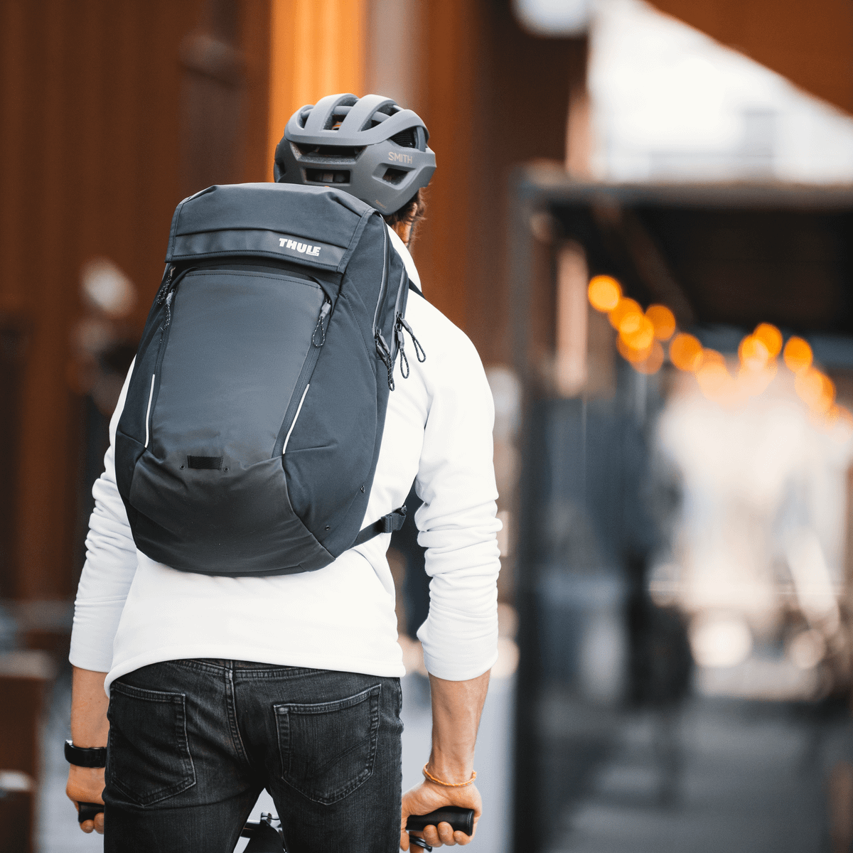 A cyclist bikes through a city street carrying a black 27L Thule Paramount Commuter Backpack.