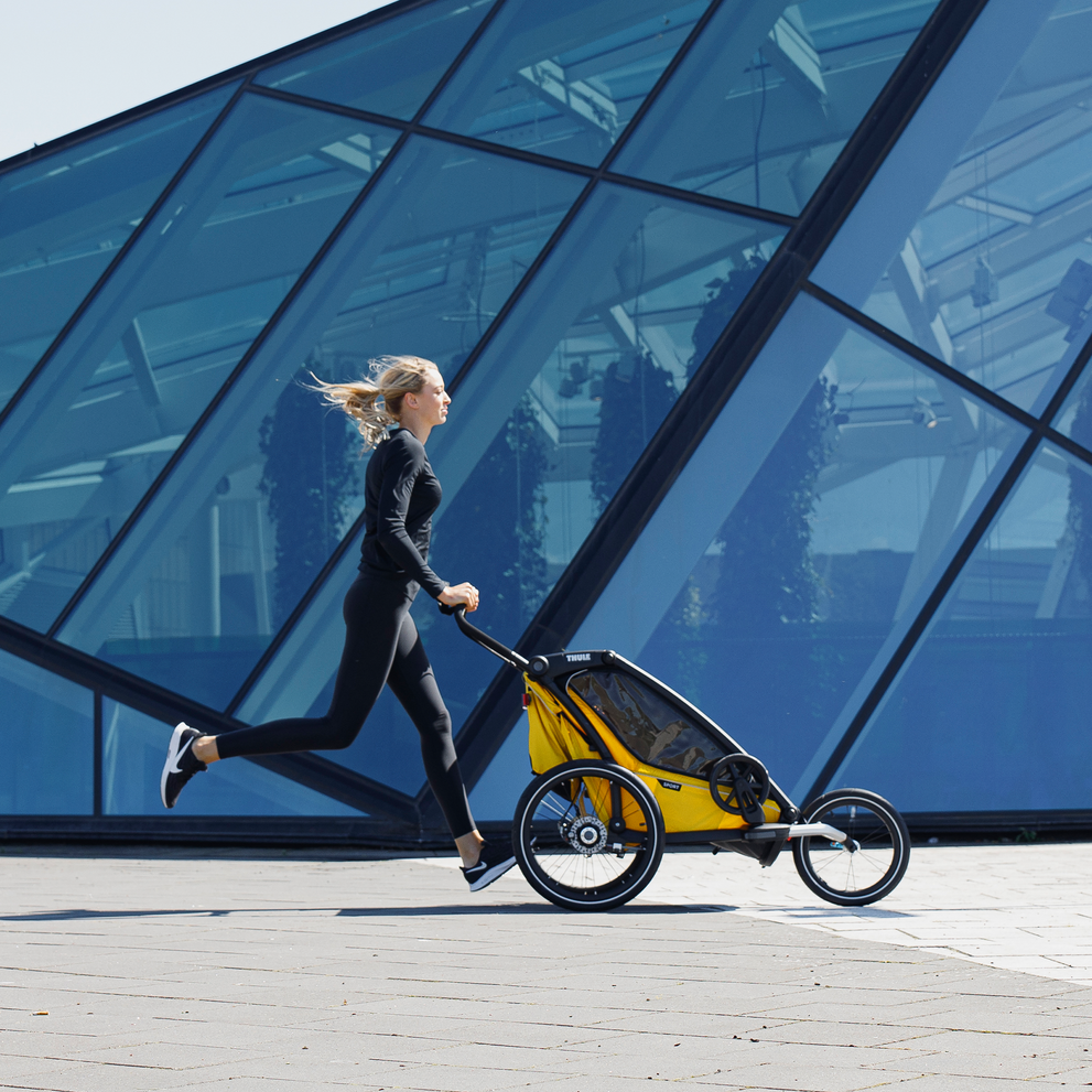 A woman ties her shoe laces beside a Thule Chariot Sport child bike trailer.