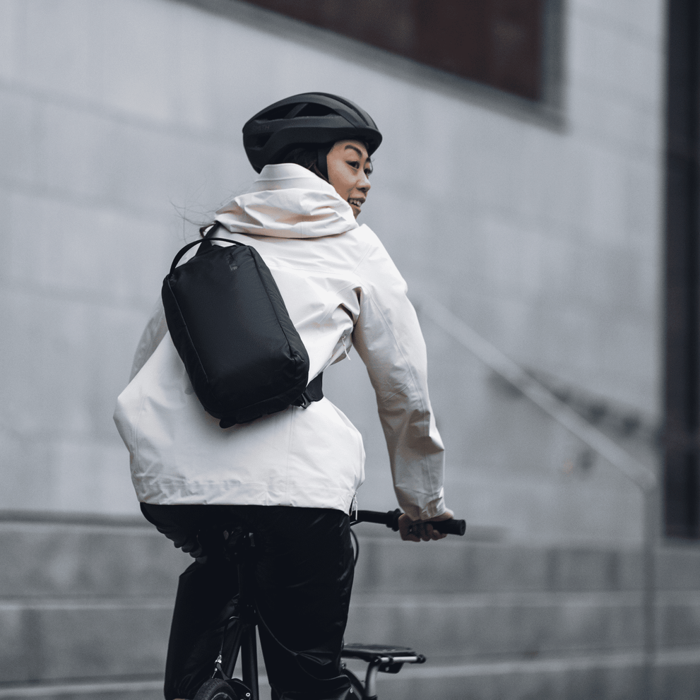 A woman bikes down a street, with a black Thule Tact Sling bag and a helmet.