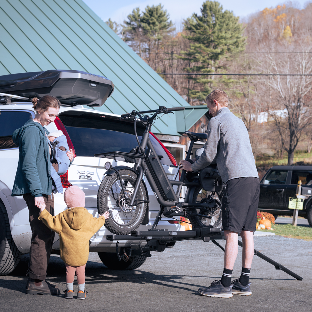 A man is securing an e-bike on the Thule Vero hitch bike rack mounted on the back of a car, parked in an outdoor setting with a woman and two smaller children next to the car.