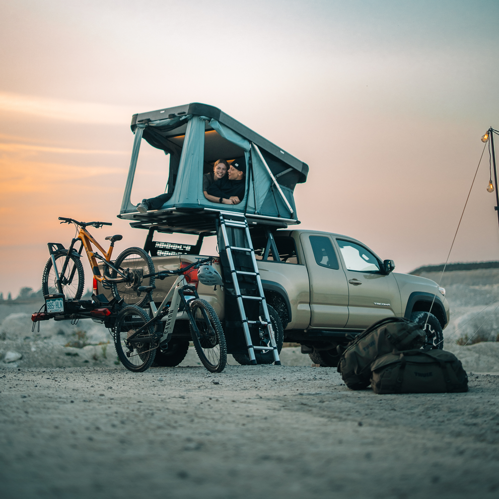 Thule Widesky hardshell rooftop tent unfolded and mounted on the top of a car with two bikes on the towbar bike rack, parked in an outdoor setting with two people inside the rooftop tent.