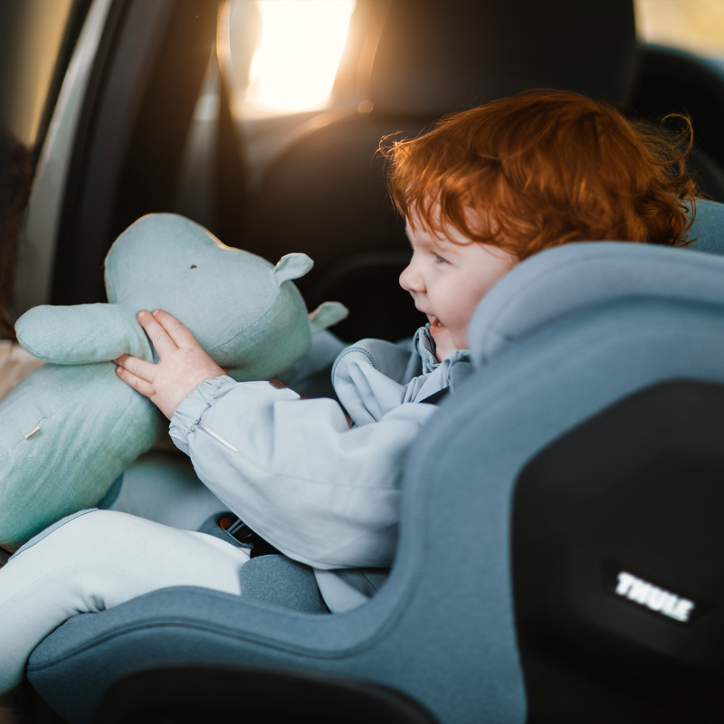 An orange-haired child smiling and holding a plush toy while sitting in a Thule Elm toddler car seat.