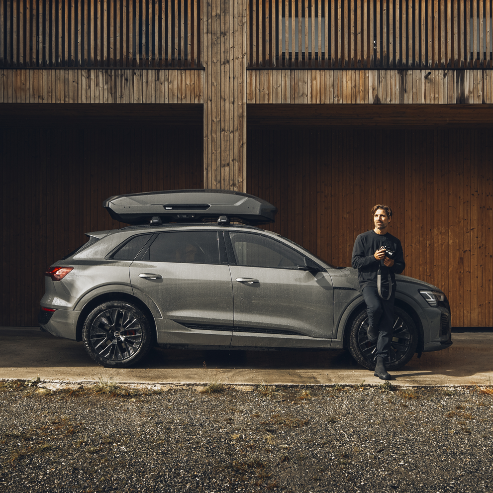 A man leans against a vehicle with a Thule Motion 3 roof box parked next to a wooden wall.