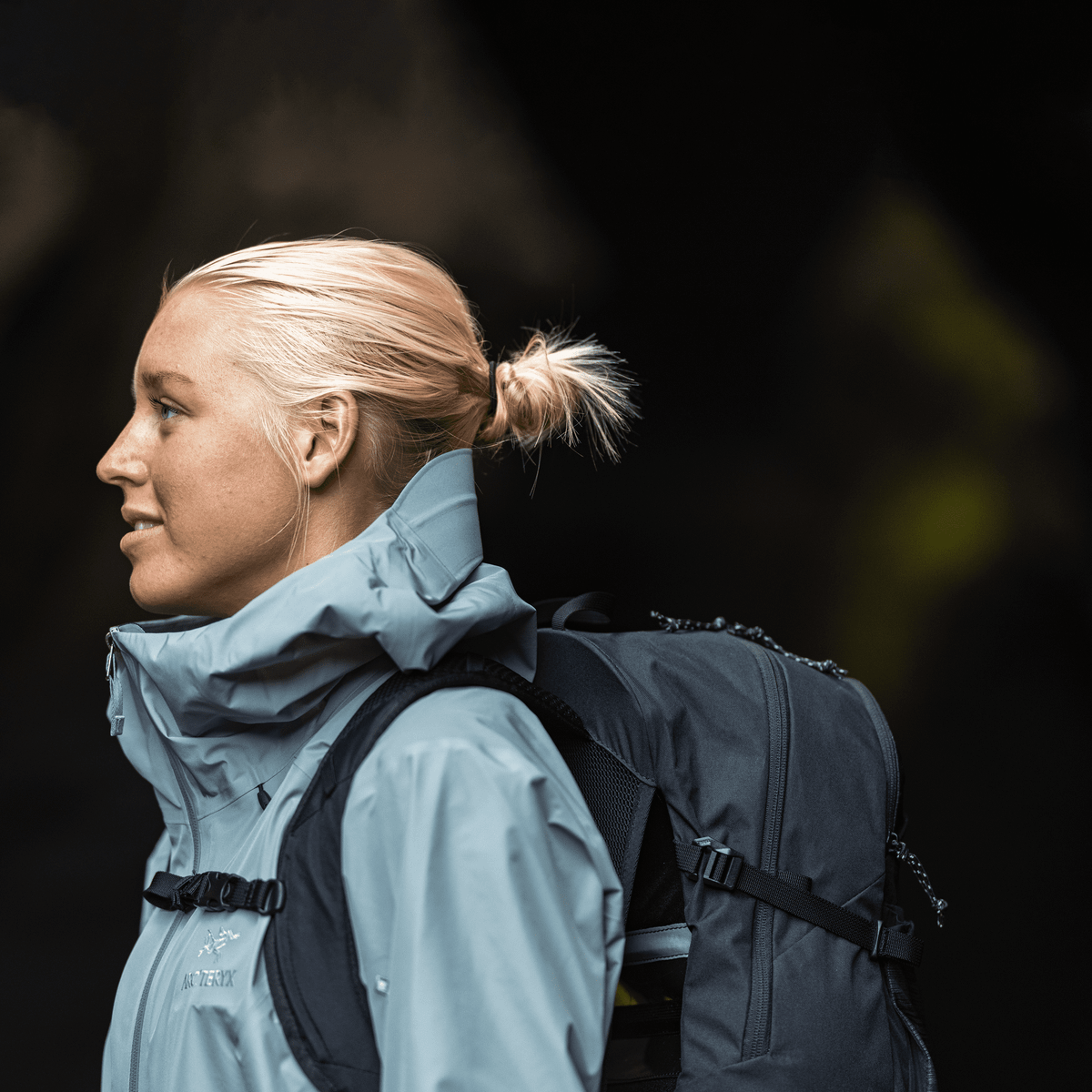 A close up of a blonde woman with a blue jacket carrying a black Thule Nanum 18L hiking backpack.