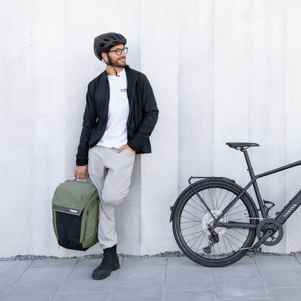 A man with a beard and glasses, dressed in a black jacket and beige pants, stands beside a bicycle holding a green Thule bag, against a concrete wall.