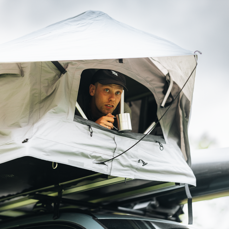 Thule Foothill 2 soft-shell rooftop tent unfolded and mounted on top of a car with a man inside the tent drinking coffee.