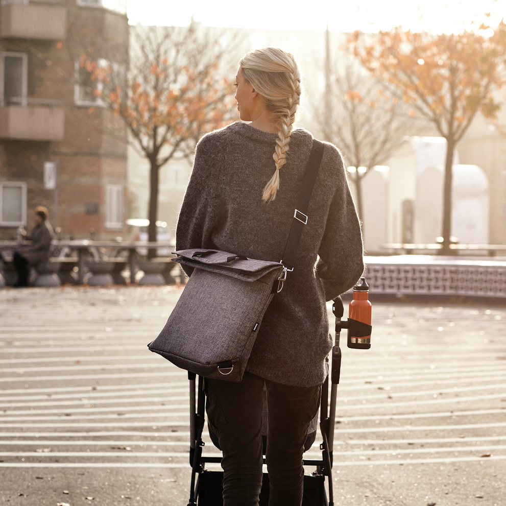 A woman walks through a park with a stroller and a gray Thule Changing Bag around her back.