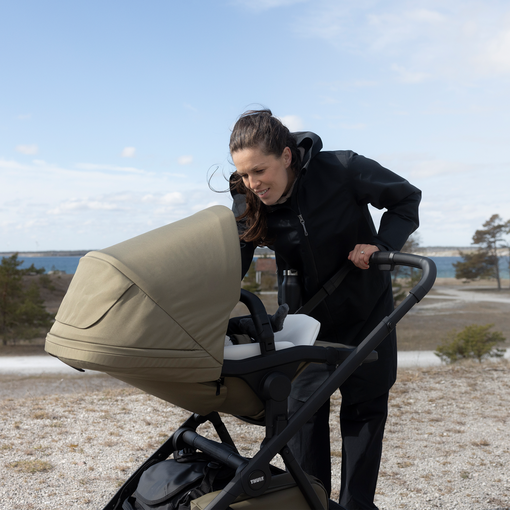 Adult checking on a baby in a Thule Charm stroller bassinet outdoors by the water.