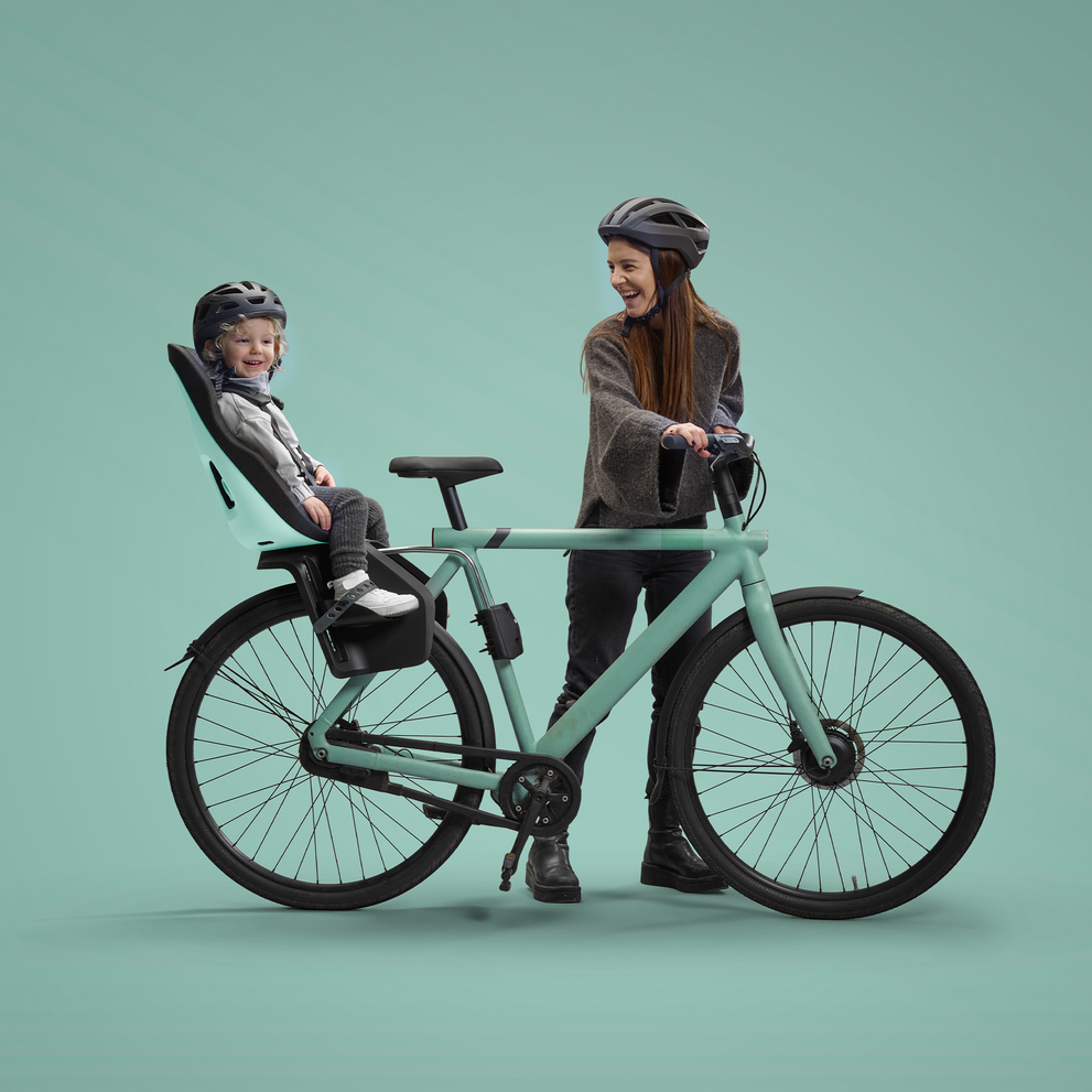 A cheerful child wearing a helmet sits in a rear-mounted child's bike seat on a pastel green bicycle. A woman, also in a helmet, stands astride the bicycle, smiling down at the child