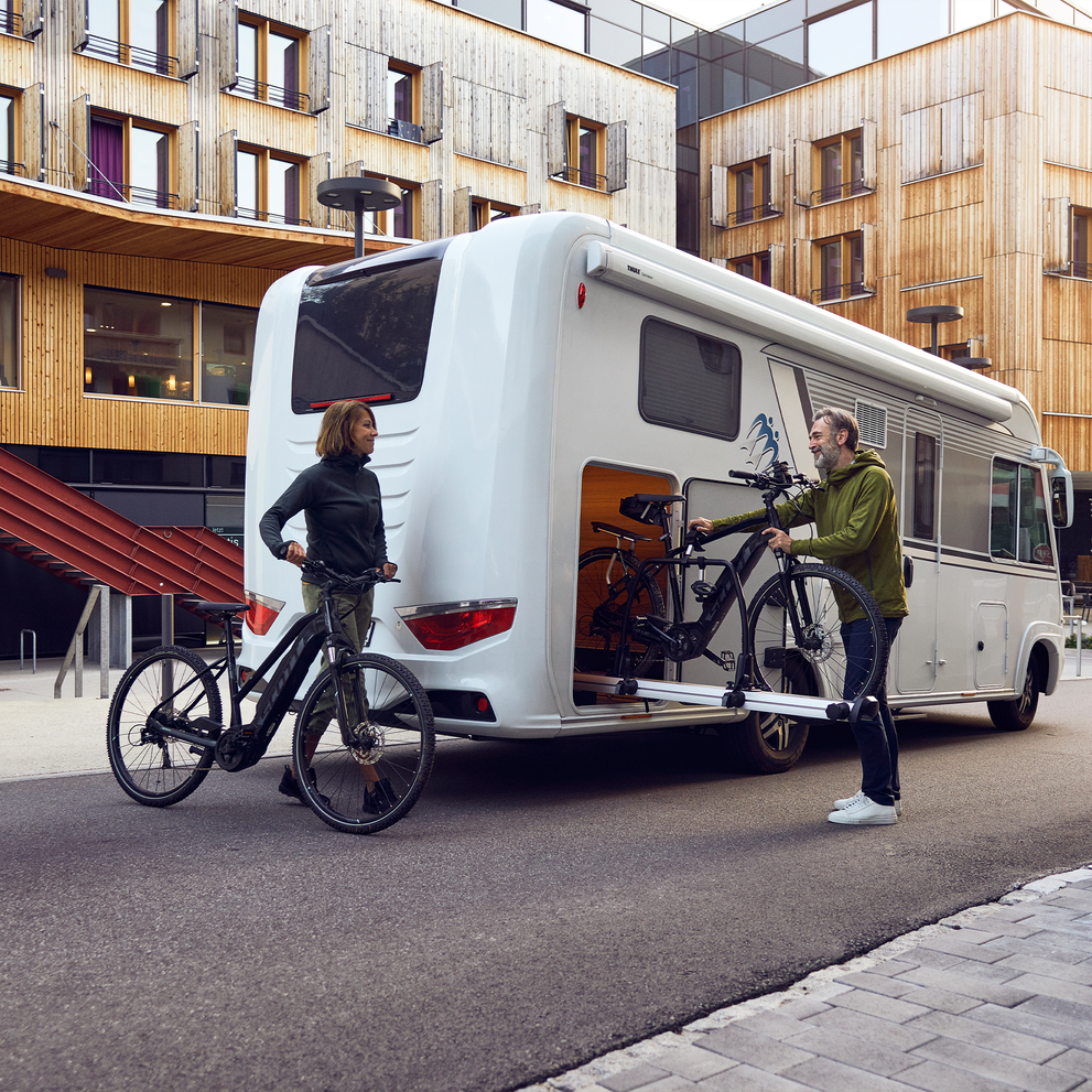 An rv is parked in the city and a couple takes their bikes out of a Thule Veloslide Standard bike rack.