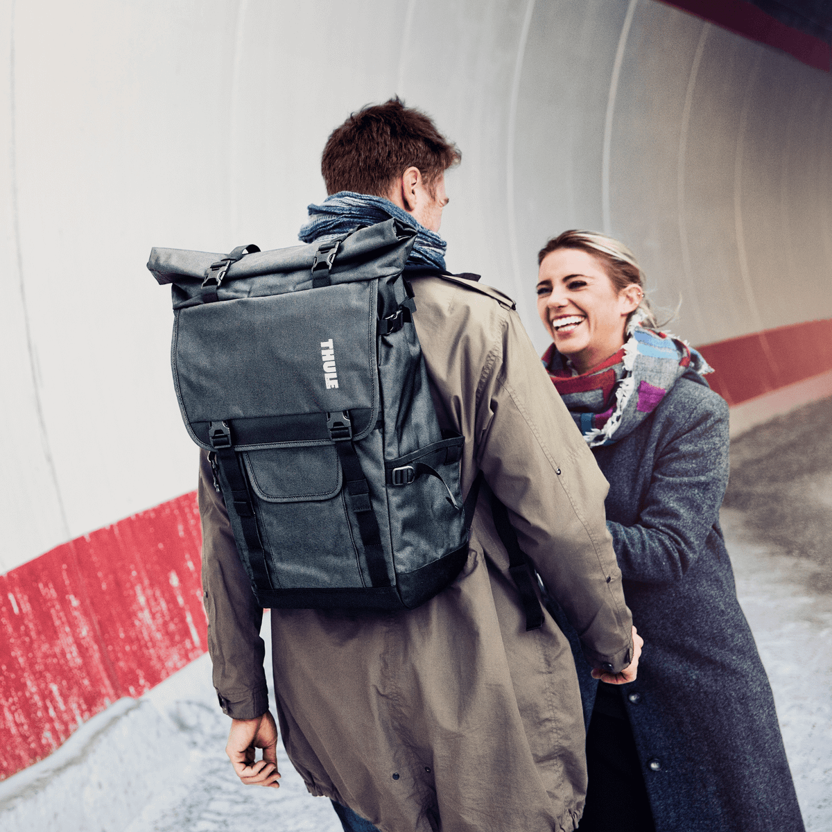 A man speaks to a woman next to a concreate wall carrying a Thule Covert DSLR Rolltop backpack.