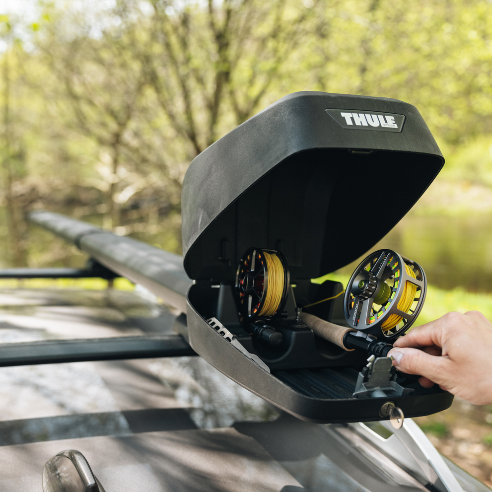 A closeup of an open Thule RodVault 2 fly rod carrier installed on the roof of a car in a forest setting.