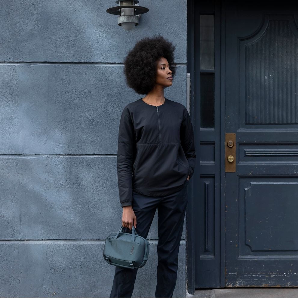A woman stands by a blue wall holding a Thule Subterra Crossbody 5L.