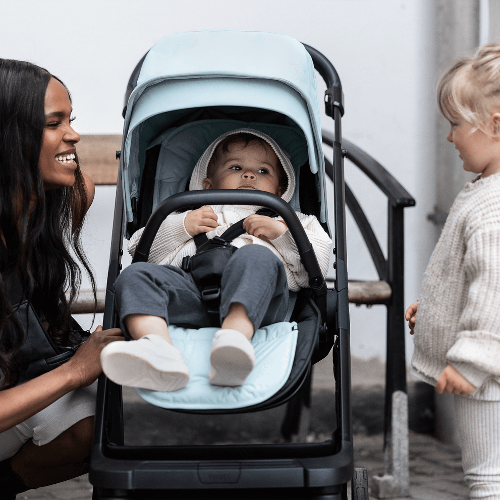 A woman smiles at a toddler and kneels next to her baby in a stroller with a blue Thule Stroller Seat Liner.