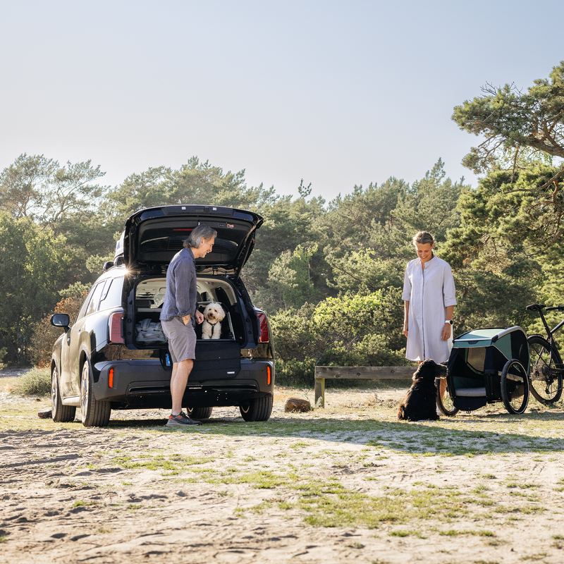 Man standing by an SUV with a white dog inside a Thule Allax dog crate, while a woman stands nearby with a black dog and a bicycle trailer in a scenic forest setting.