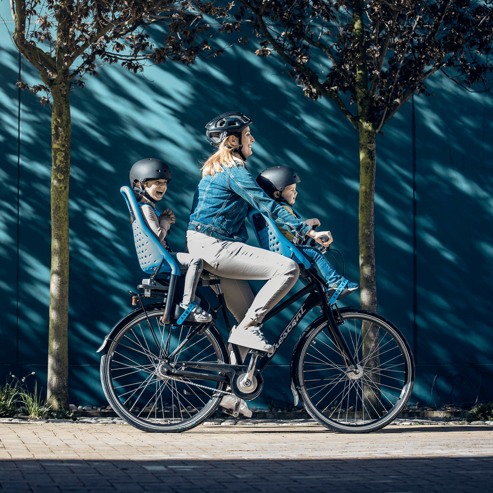 A woman bikes down a street with a blue wall, with her kids in a rear and front Thule Yepp Maxi child bike seat.