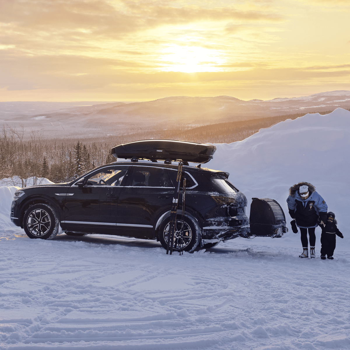 A parent with child unloading their ski gear from a car with a roof top box in a snowy landscape