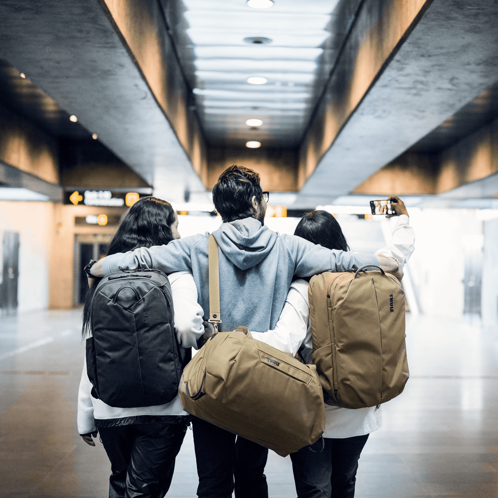 Three friends take a selfie in a metro station with backpacks and a tan Thule Aion Duffel.