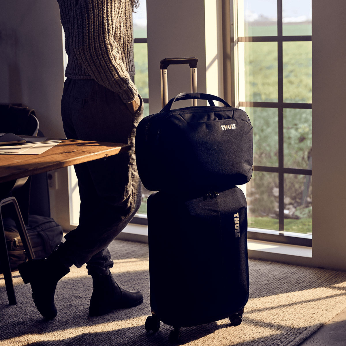 A woman looks outside a window at the sunshine with backpack on her Thule Subterra Carry-On Spinner suitcase.