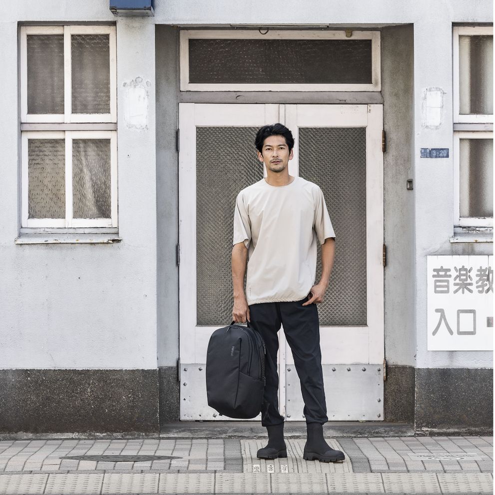 A man stands outside a white door in a t-shirt and black trousers holding a Thule Subterra backpack.