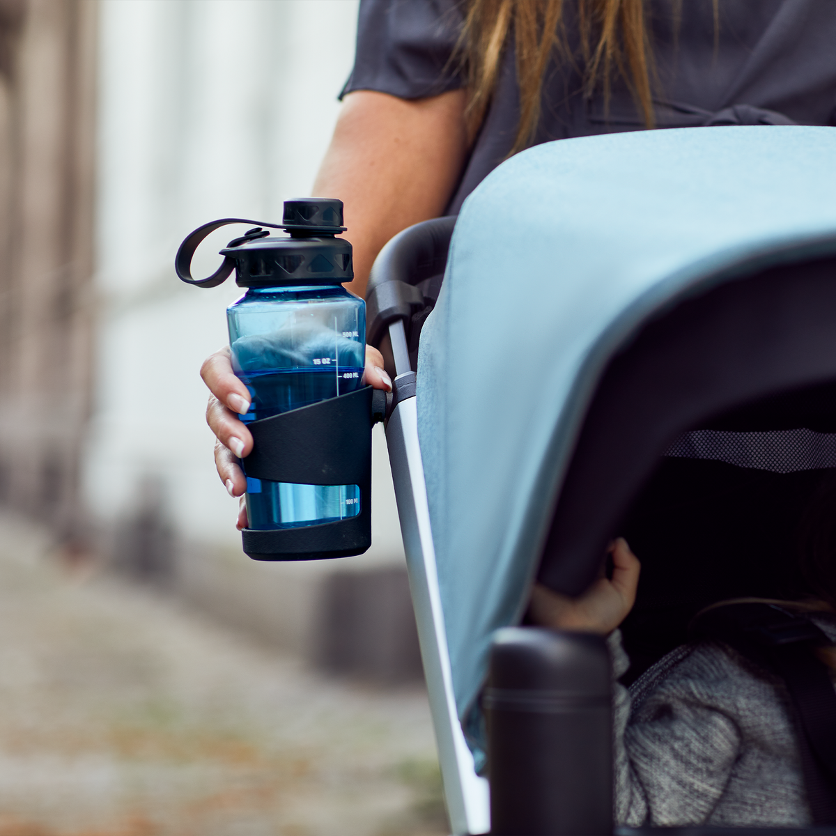 A toddler sits in a blue stroller with a Thule Stroller Cup Holder with a blue water bottle.