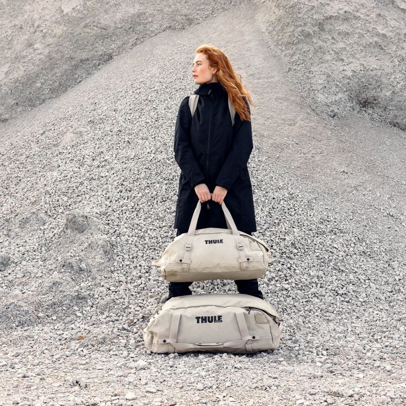 A woman with long red hair stands in a rugged outdoor environment, holding a large duffel bag with both hands as she gazes into the horizon. A second sand-colored duffel bag lies at her feet.