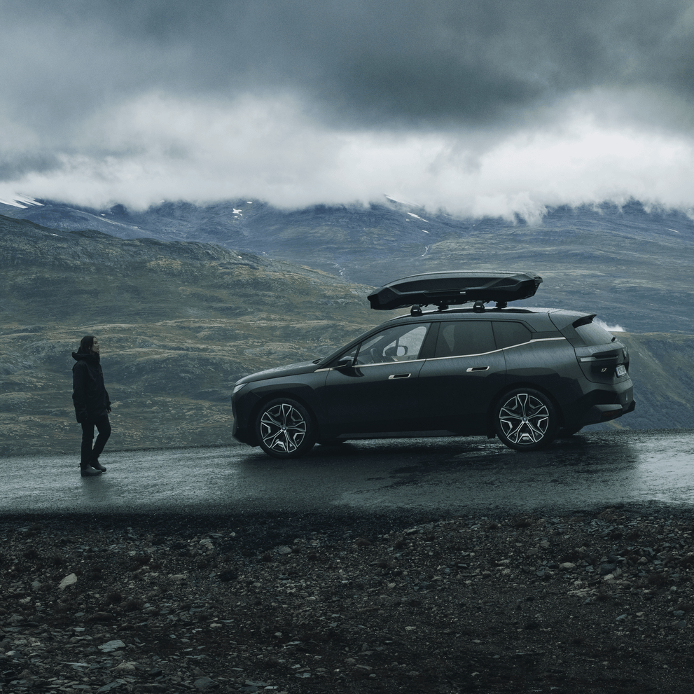 A person walks towards a vehicle parked next to the mountains with a black Thule Motion 3 roof box.