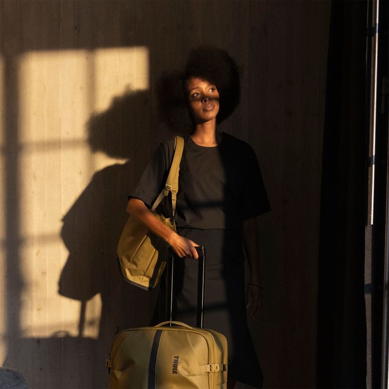 Femme près d'une fenêtre avec une valise roulante Thule Aion sous un soleil chaleureux.