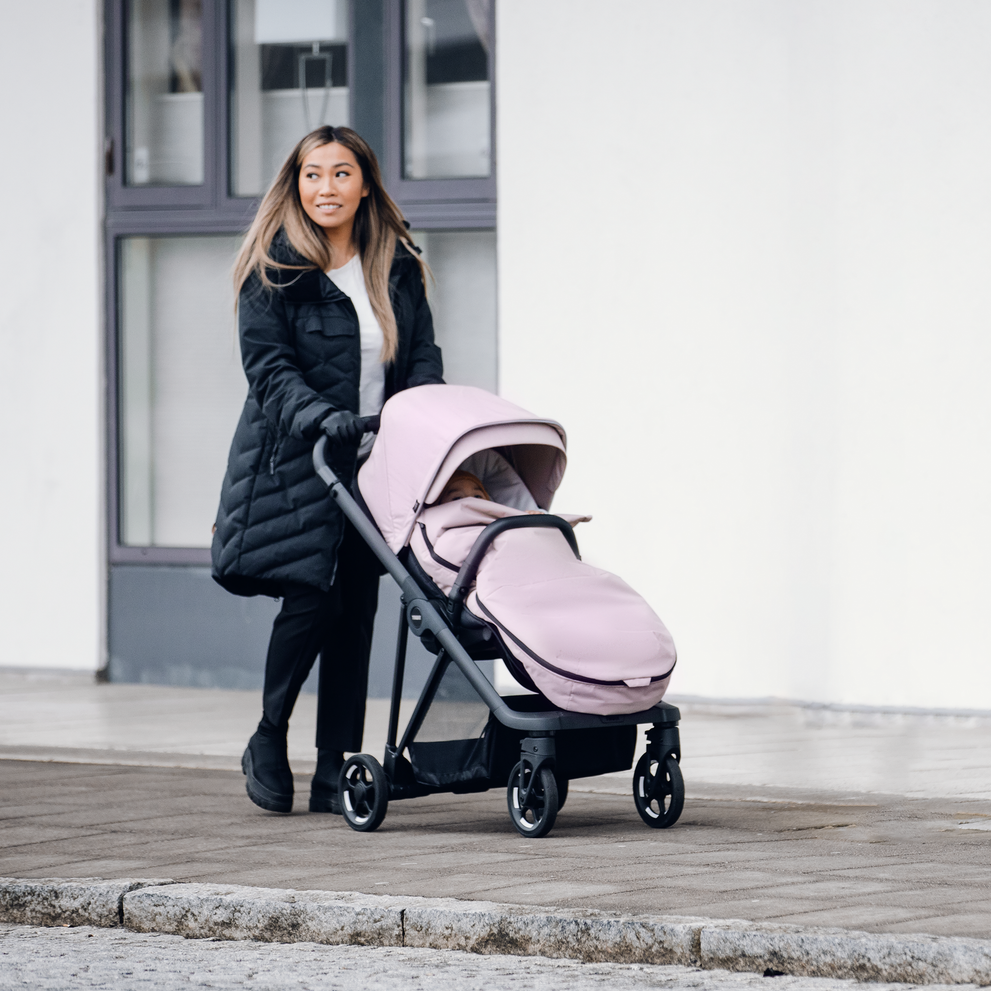 A woman in a jacket walks with her stroller and her baby in a pink Thule Stroller Footmuff.