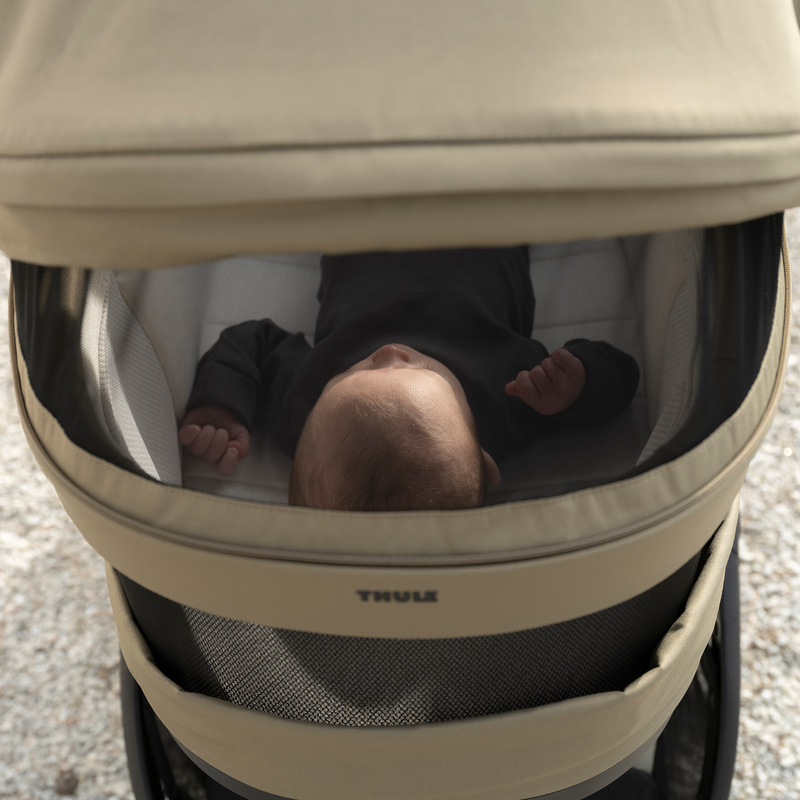 Baby lying inside a Thule Charm stroller bassinet with a mesh cover partially closed.