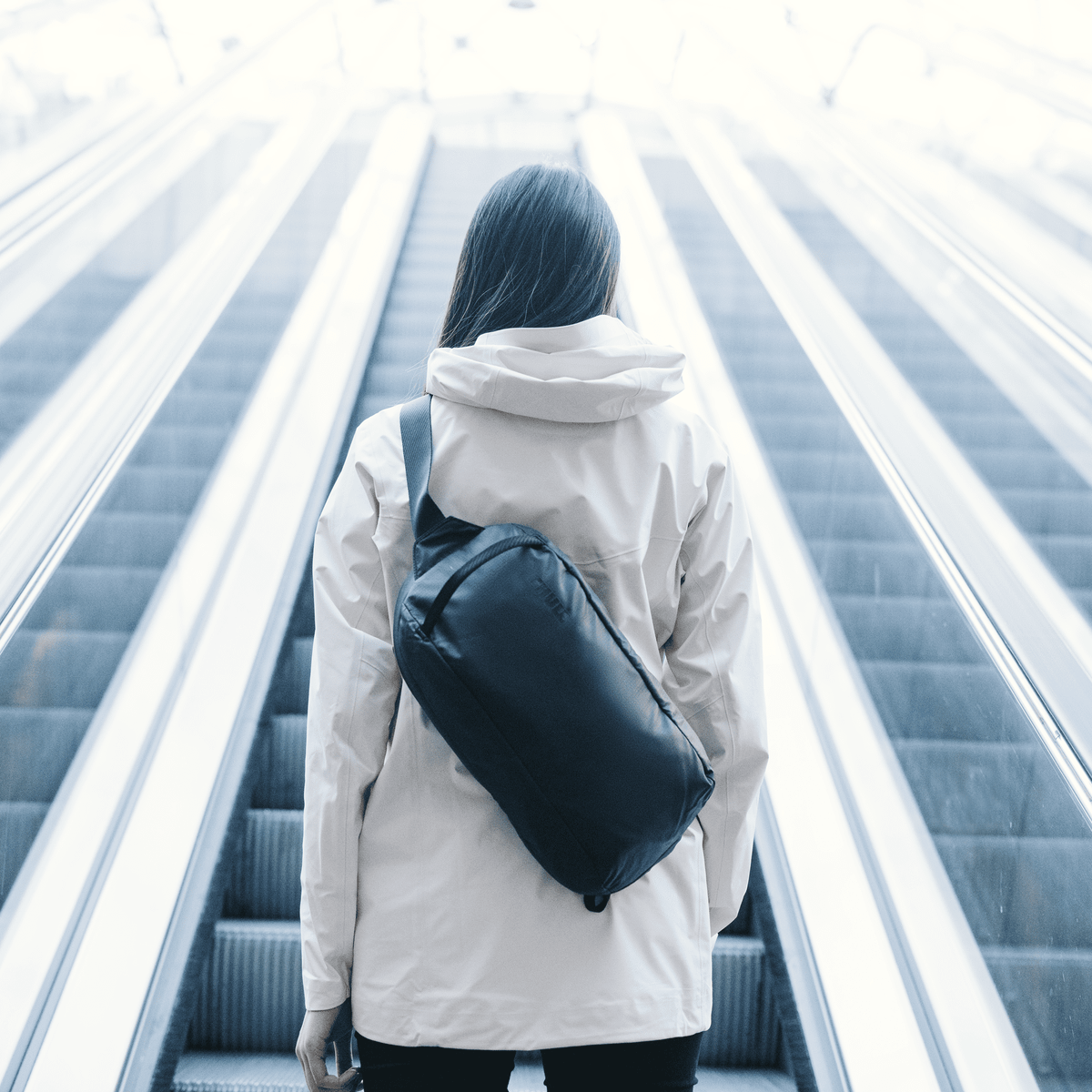 A woman goes up escalators into the sunshine, with a white jacket and a Thule Tact Sling bag.